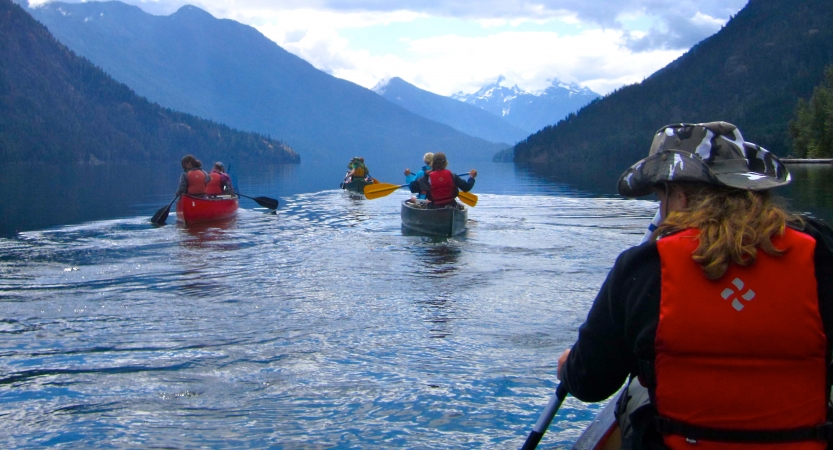 a group of outward bound students paddle canoes on calm water surrounded blue mountains